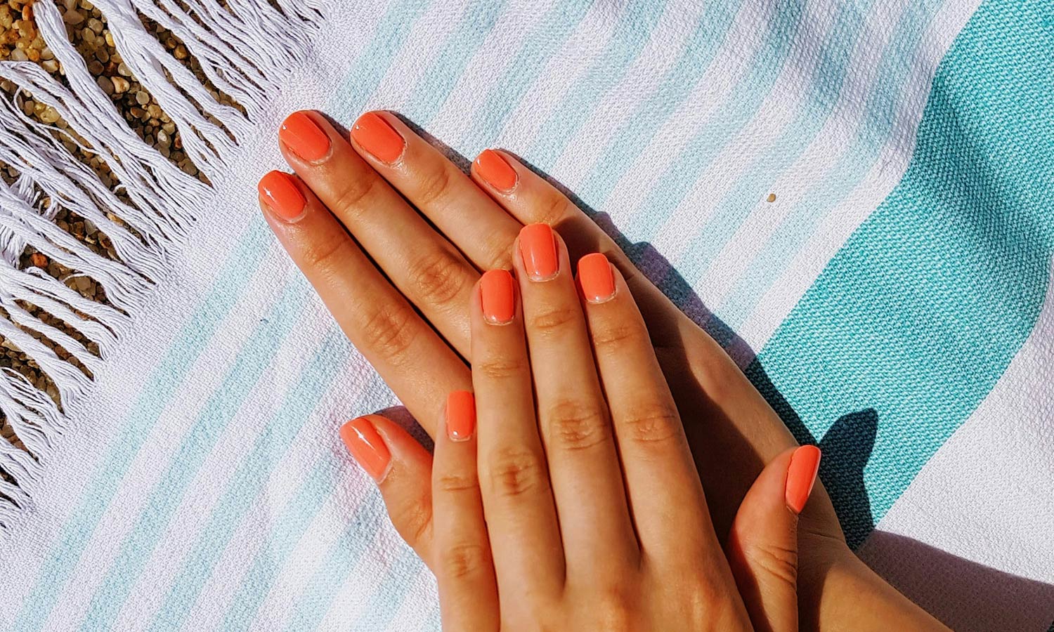 coral semi-cured gel nails on hands on top of a blue and white stripped beach towel at the beach