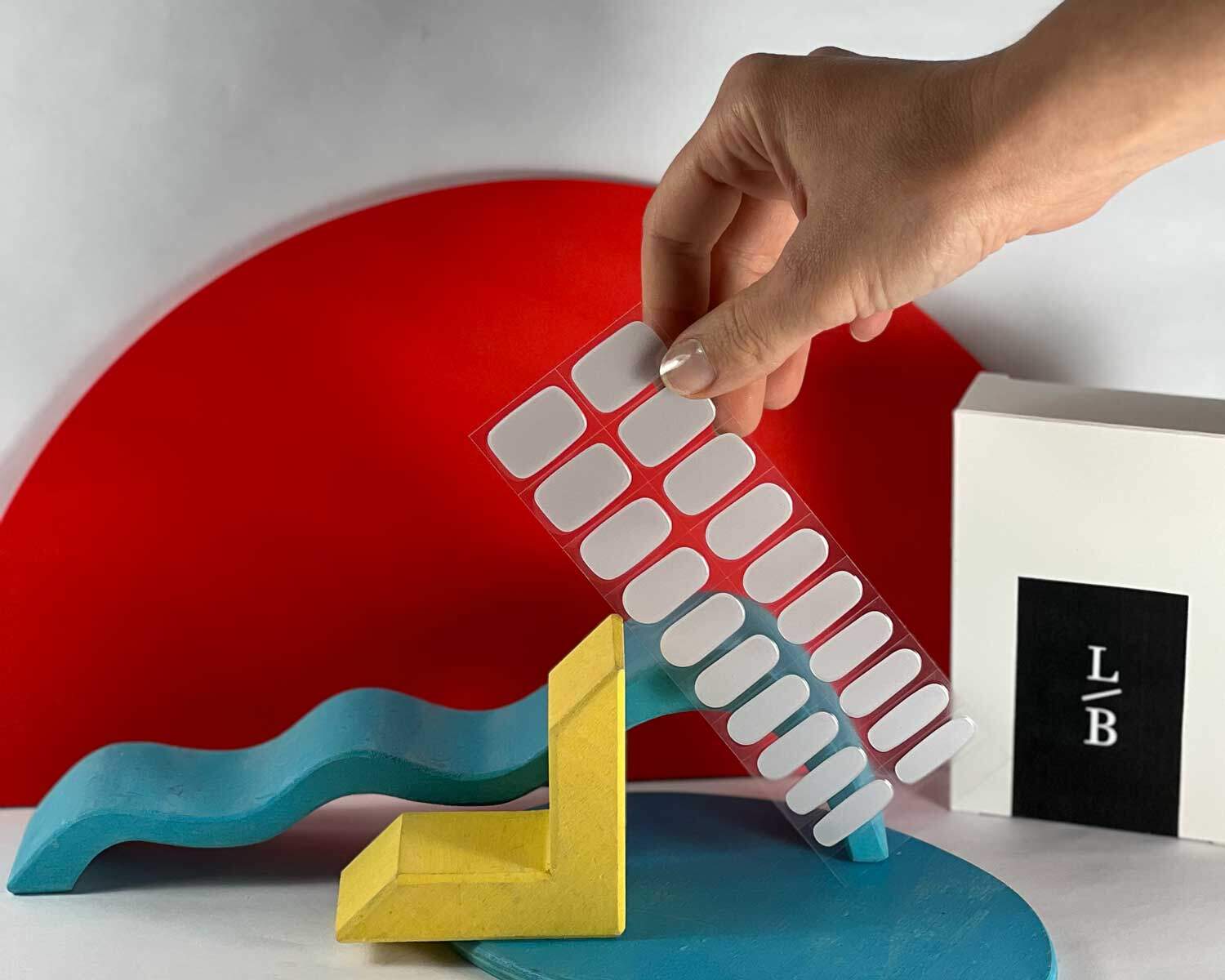 Hand holding Grey diamond silver semi-cured gel nails in mockup to show product against blue, red and yellow wooden blocks with la-bek uv lamp in the background