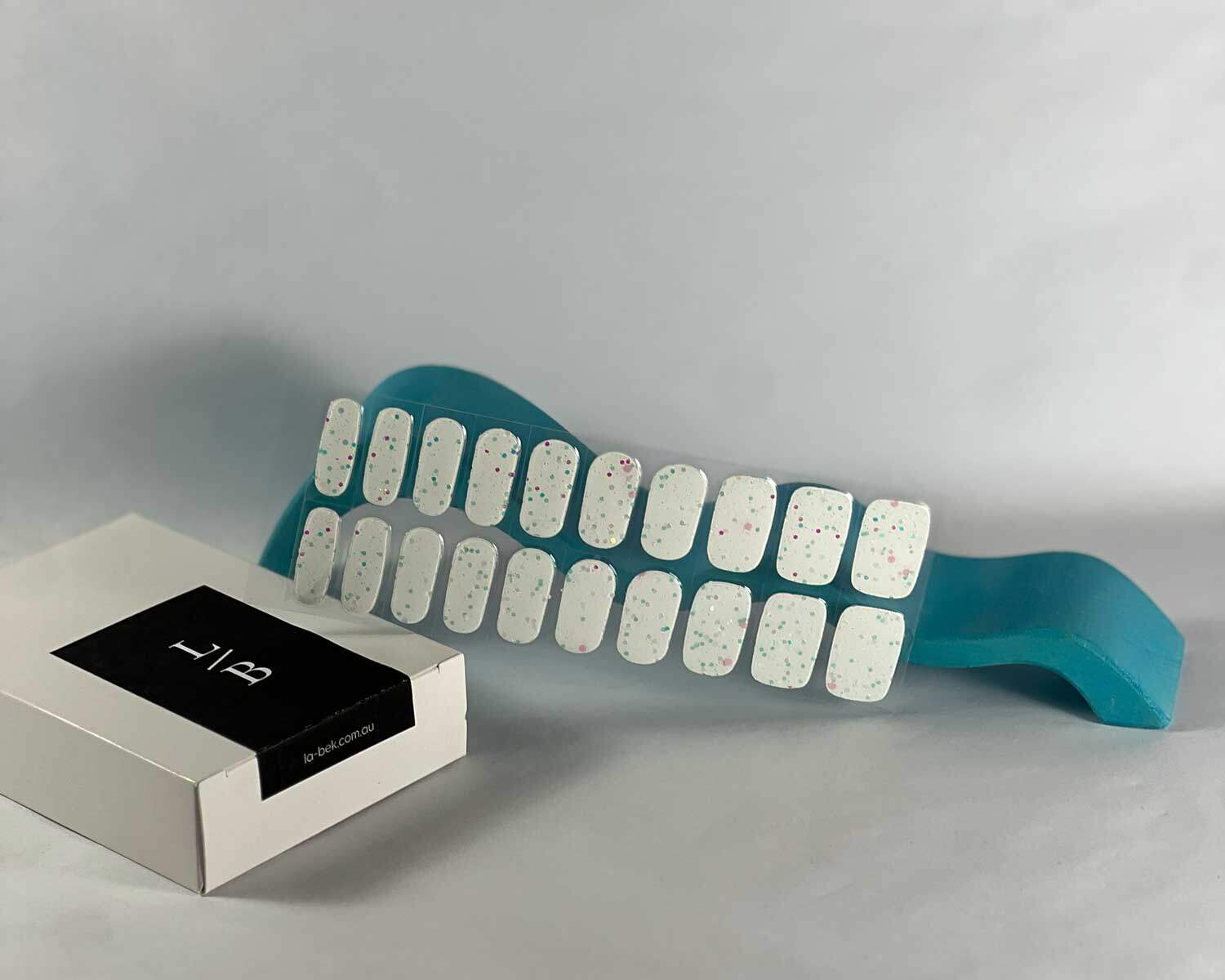 Sprinkle snow Glow in the dark white semi-cured gel nails in mockup to show product against blue wave wooden block with la-bek uv lamp in the background