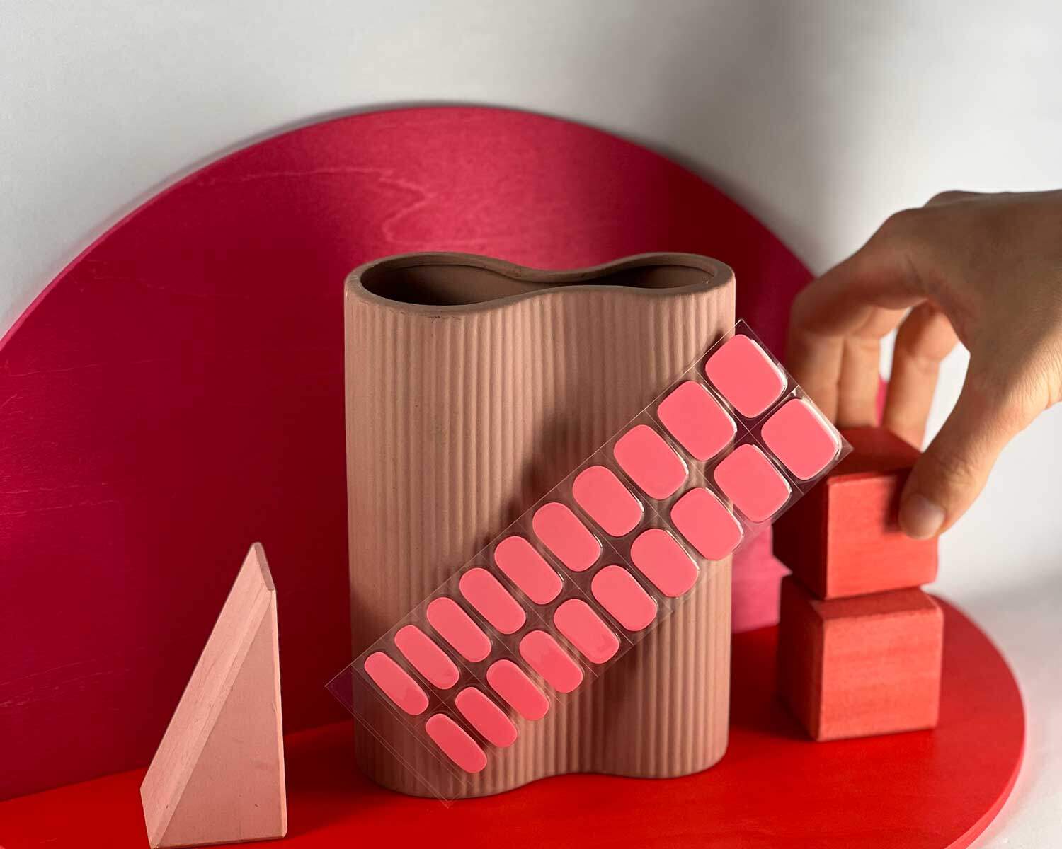 Watermelon pink semi-cured gel nails in mockup to show product against red and pink wooden blocks with hand touching blocks